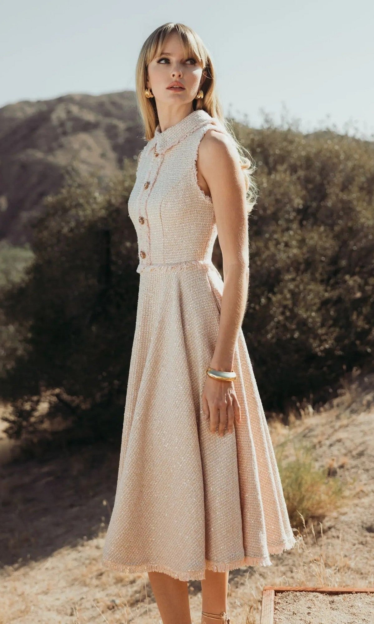A woman stands outdoors against hills and trees, wearing the blush Mac Duggal 27658 Semi-Formal Midi Dress, styled sleeveless with bracelets and earrings.